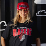 Person wearing a black Portland Trail Blazers t-shirt and red cap against a black and white striped background with cloud shapes.