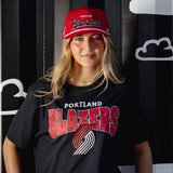 Person wearing a black Portland Trail Blazers t-shirt and red cap against a black and white striped background with cloud shapes.