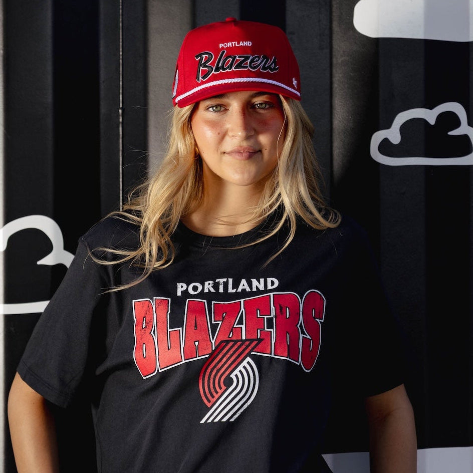 Person wearing a black Portland Trail Blazers t-shirt and red cap against a black and white striped background with cloud shapes.