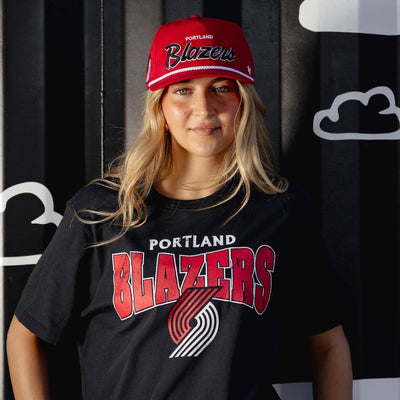 Person wearing a black Portland Trail Blazers t-shirt and red cap against a black and white striped background with cloud shapes.
