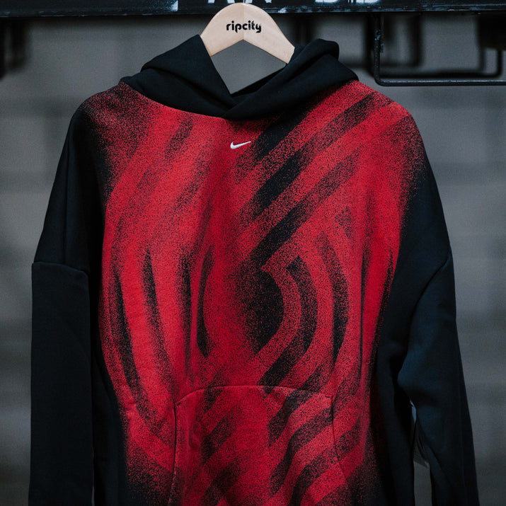 A black and red patterned Nike hoodie hanging on a wooden 'rippity' hanger against a dark gray brick wall background.