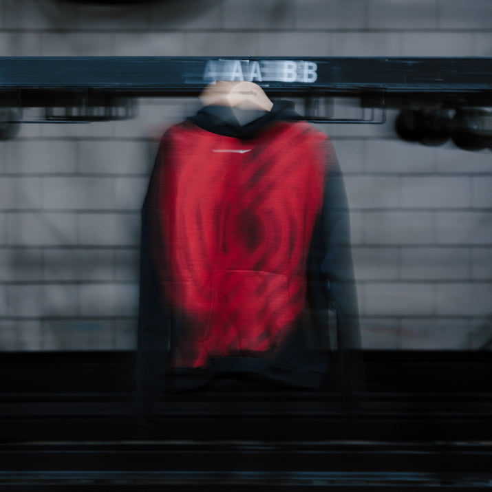 A red and black hooded sweatshirt is displayed on a hanger inside a dark industrial rack or storage unit with blurred horizontal motion lines across the frame.