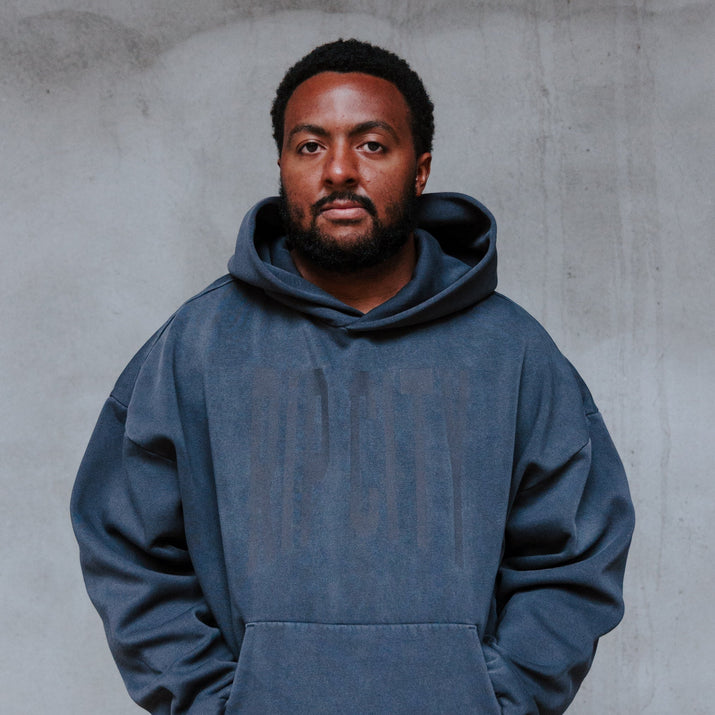 A man wearing an oversized slate blue hoodie with a tonal 'RIP CITY' graphic on the chest, standing against a textured gray concrete wall.