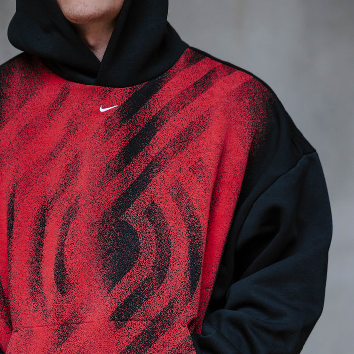 A close-up of a person wearing a Nike pullover hoodie with a red-and-black abstract-patterned Trail Blazer pinwheel logo front and solid black sleeves, hood, and lower body. A small white Nike swoosh is visible on the chest.
