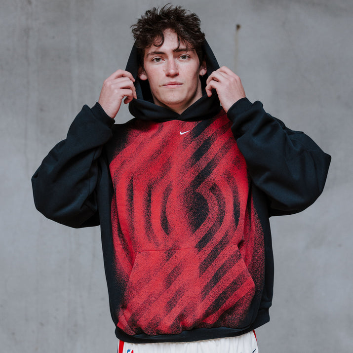 A young man wearing a black and red patterned hoodie with white basketball shorts, posing against a textured gray concrete wall.