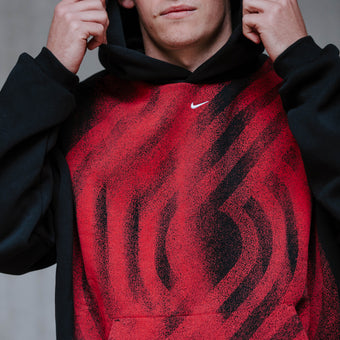 A person wearing a red-and-black Nike hoodie with a textured, wavy pattern on the front, holding the hood up against a gray concrete wall. Blazer Pinwheel design is on front.