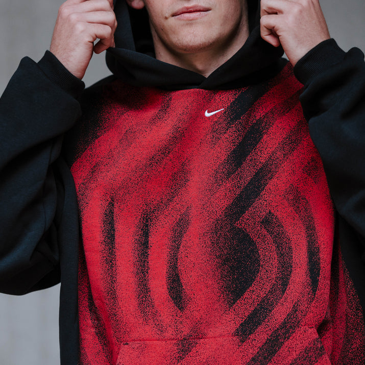 A person wearing a red-and-black Nike hoodie with a textured, wavy pattern on the front, holding the hood up against a gray concrete wall. Blazer Pinwheel design is on front.