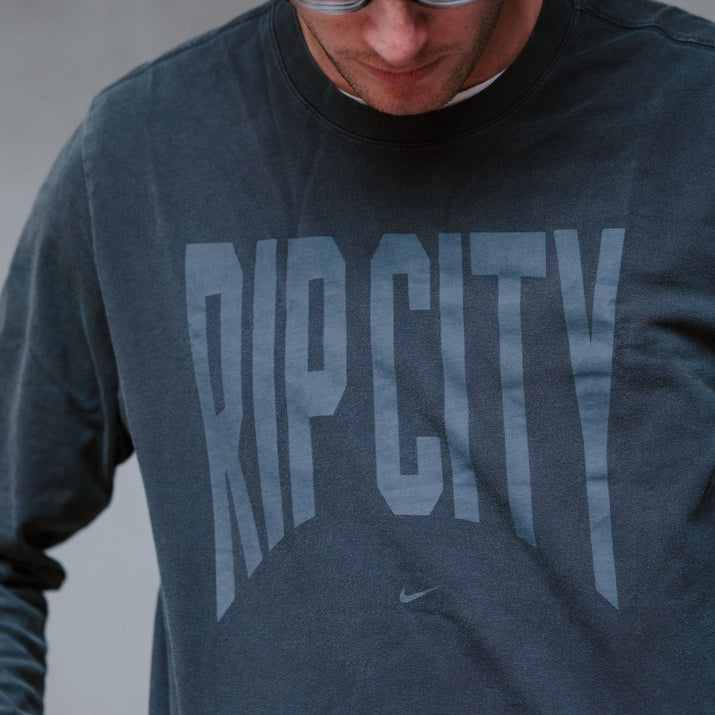A close-up of a person wearing a dark charcoal gray long-sleeve crewneck sweatshirt with 'RIP CITY' printed in a faded gray font across the chest. A small Nike swoosh is visible below the text.