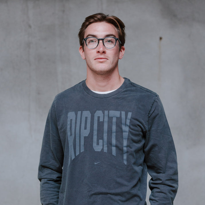 A man wearing a dark gray long-sleeve t-shirt with 'RIP CITY' printed across the chest in a faded gray font, paired with black pants, standing against a textured gray concrete wall.