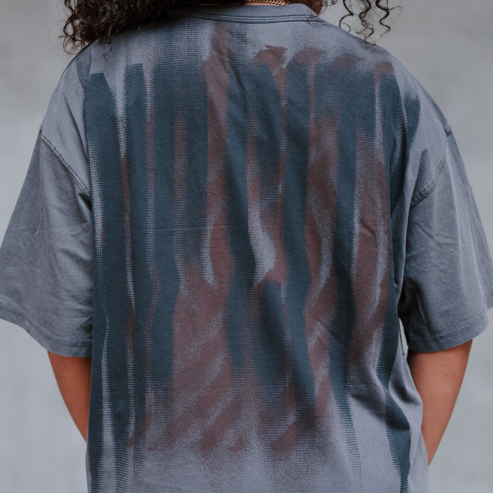 A person viewed from behind wearing an oversized short-sleeve T-shirt with a faded Rip City spray paint pattern in slate blue and muted brown on a light gray background.