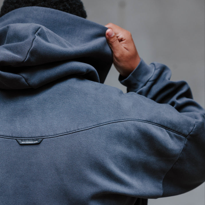 A person viewed from behind wearing a dark slate blue hooded sweatshirt, holding up the hood with both hands against a textured gray concrete wall.