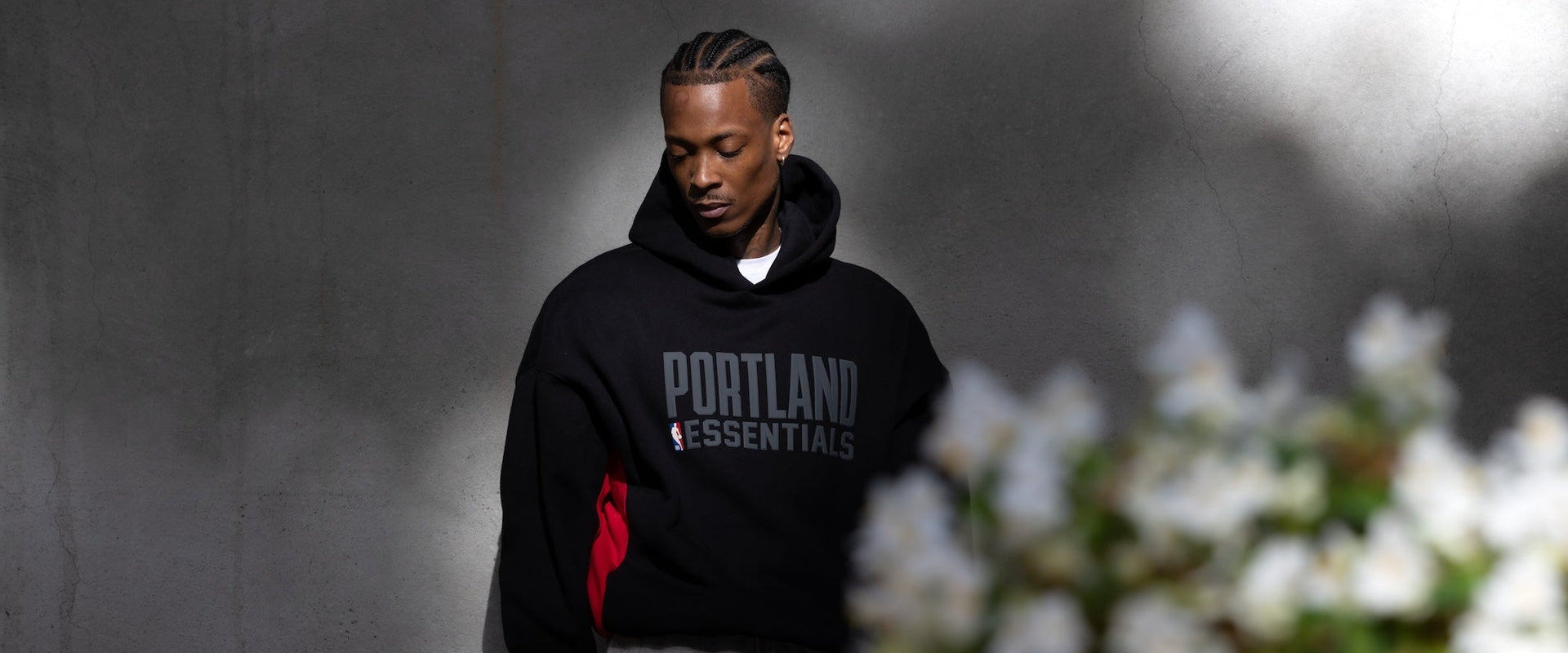A man wearing a black Portland Essentials hoodie with a red side accent and gray sweatpants, standing against a gray concrete wall with blurred white flowers in the foreground.