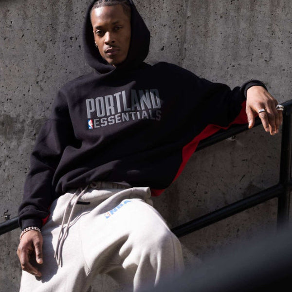 A man wearing a black Portland Essentials hoodie with white text and a red side panel, paired with light beige sweatpants, sitting on a black metal staircase against a concrete wall.